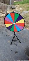 Photo showing the multi-colored spinning wheel fully visible on a black tripod stand outdoors on pavement next to a grassy area.