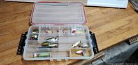 Open Plano tackle box showing various fishing lures inside compartments on wooden surface