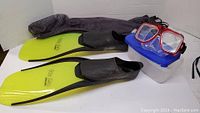 Photo showing the lime green Rip Tide flippers, red snorkel mask placed on top of a clear container with a blue lid, and a gray mesh carrying bag behind them.