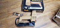 Beige and black Swingline electric nailgun displayed on floor with unopened nail strip and original box in the background.