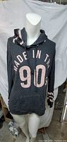 Front view of grey women's hoodie with 'Made in the 90s' printed in large pink letters and numbers, displayed on mannequin.
