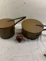 Full view of the two antique copper pots with lids alongside an apple for size comparison, showing the pots' proportions and handles.