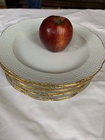 Stack of ten white porcelain dinner plates with textured rims and gold trim, apple placed on top for scale.