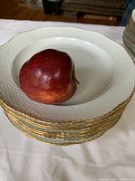 Stack of ten white porcelain dinner plates with visible textured pattern and gold trim edges. A red apple placed on top for scale shows plates are approximately 9.5 inches in diameter.
