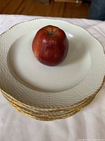 Top view of stack of ten white KPM porcelain dinner plates with an apple on top for scale, showing the embossed lattice border and gold trim.