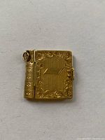 Front view of small gold-tone book-shaped locket showing decorative detailed engravings and hinge with a small loop for a chain.