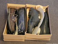 Two boxes side by side, each with one pair of men's shoes: brown boat shoes in one, black dress shoes in the other. Both appear new or nearly new.