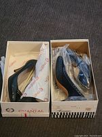 Top-down view of two boxes containing one pair each of black vintage designer heels from Chantal and Aguane, wrapped in branded tissue paper and plastic.