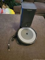 Photo showing iRobot Roomba i4 robot vacuum placed in front of the Clean Base automatic dirt disposal station with power cord visible.