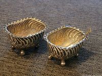 Two small antique sterling silver salt cellars placed side by side on fabric surface showing embossed scrolling and floral detailing, scalloped rims and ball feet.