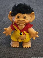 Front view of vintage troll doll with tan body, large ears, black hair, and red-yellow outfit.