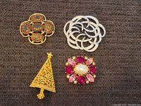 Four brooches arranged on fabric: a gold-tone floral enamel brooch in oval shapes, a silver-tone crescent linked brooch, a rhinestone-studded Christmas tree brooch, and a pearl and ruby-like rhinestone floral cluster brooch.