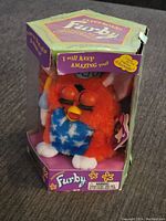 Front angled view of vintage Furby inside original hexagonal box, showing orange fur with blue star pattern and closed eyes. Box edges show wear and tear.