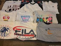 Photo showing ten assorted vintage graphic shirts laid out including La Jolla, FILA, California themed prints, and a crown logo.