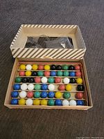Top perspective of open vintage cardboard box filled with several rows of solid color glass marbles arranged by color.