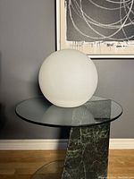 Large white plastic spherical LED ball lamp seen resting on a glass and marble top table from side angle.