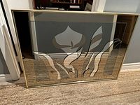 Frontal view of mid century modern art glass mirror with floral design, in a gold metallic frame, placed on floor leaning against a wall.