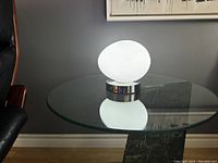 Close-up image of the modern egg-shaped lamp illuminated on a glass side table against a gray wall.