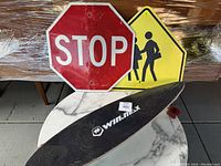 Skateboard with black deck and red wheels displayed with stop and school crossing street signs on white marble surface.