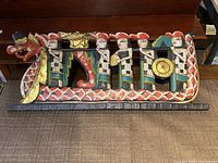 Full view of the Balinese wooden plaque depicting a dragon-like creature carrying multiple human figures sitting along the back, painted in vibrant colors.