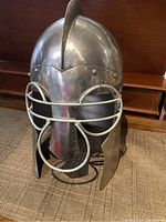 Front view of a shiny silver-toned metal helmet with a raised crest and white metal face guard bars.