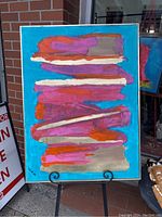 Front view of the original abstract painting featuring layered strokes of pink, orange, beige, and white on a blue background, displayed on an easel outside a storefront.