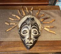 Front view of vintage African wooden mask on a wooden surface showing carved facial features and attached bamboo sticks radiating outward.