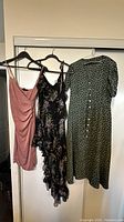 Photo of three size Small dresses hanging on black plastic hangers against a white wall and window. Pink ruched sleeveless dress, black floral spaghetti strap dress with ruffles, green polka dot dress with short sleeves and buttons, one button missing on green dress.