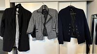 Five women's blazers hung on hangers against a white closet door showing various colors and styles: black, gray with trim, navy blue with metal buttons.