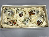 Box containing six cream-colored double-sided glass ornaments with detailed Dickens' Christmas Carol scenes, cushioning tissue around each.