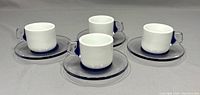 Four white ceramic espresso cups with blue accents on handles and bases, each on a clear glass saucer.