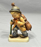 Front angle view of the Hummel Little Cellist figurine showing detailed painting and facial expression, with walking stick and cello case