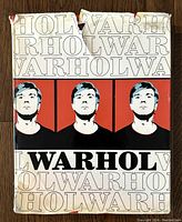 Front view of the dust jacket showing three stylized portraits of Andy Warhol and repeated text 'WARHOL'. Upper edge shows damage and rips.