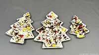 Three ceramic Christmas tree shaped trays arranged in descending size order, showing splatter paint decoration in yellow, red, black, and brown on white base, on a gray background.