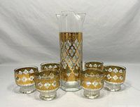 Front view showing full set of one tall pitcher and six matching short glasses with gold Valencia pattern.