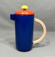 Front view of vintage Memphis Milano French Press coffee pot showing blue body, red lid with yellow knob, and beige handle.