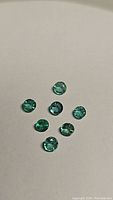 Seven small round-cut natural emeralds displayed on a neutral background, showing good luster and natural inclusions.