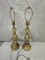 Pair of tall brass lamps side by side showing full height and shape against light backdrop.