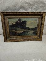 Full view of the antique oil painting and carved frame showing river landscape with trees and cloudy sky.