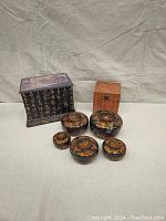 Seven decorative boxes displayed on neutral cloth background. Five circular wooden nesting boxes with floral designs in front. Two Asian-themed lacquered boxes by back. Larger black box with gold calligraphy on sides, smaller rectangular stained wood box with calligraphy.