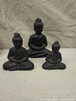 Front view of three black plaster Buddha statues of varying sizes sitting in meditation pose on a grey fabric surface with a beige backdrop.