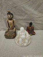 Three statues arranged together showing scale and details: white Buddha in front, wooden figure on left, red Buddha on right.
