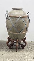 Full view of the large stoneware jar with rattan handles sitting on the carved wooden stand.