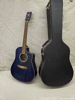 Blue Art & Lutherie Wild Cherry acoustic guitar standing next to its black hard shell case on a light background.