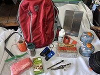 Photo showing red MEC backpack with various survival gear accessories in front of it on white fabric.