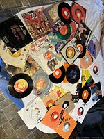 Photo showing a collection of approximately 30 45 RPM vinyl records spread out on a surface with various labels and sleeves visible.