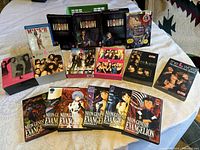 Photo shows the entire lot: 17 DVD/Blu-ray sets including The L Word various seasons, Bad Girls, Neon Genesis Evangelion series, Walt Disney's Sleeping Beauty and M.A.S.H. Blu-ray, arranged on a bed.