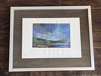 Framed artwork showing the full piece with matting and floating silver frame on wooden floor background.