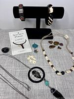 Photo showing two bracelets on display stand, multiple necklaces with beads and pendants laid out, polished stone collection, and booklet about garnet stones.