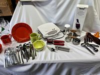 Full assortment of kitchen items showing cutlery, mugs, plates, waffle iron, mandoline box, measuring cups, spoons, wooden salad hands, and small kitchen gadgets